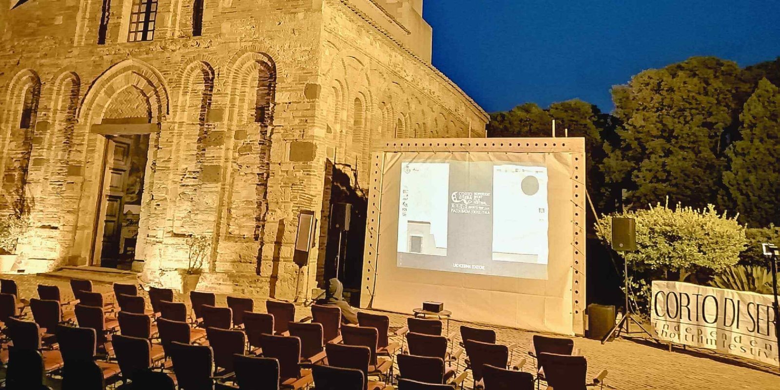 Proiezione serale all’aperto del festival Corto di Sera davanti alla Chiesa Arabo-Normanna dei Santi Pietro e Paolo a Itala, con sedie rosse vuote, schermo illuminato e cielo blu notturno.